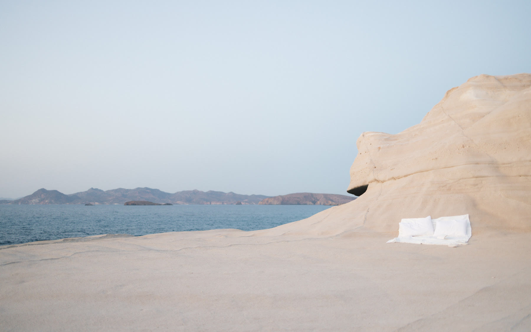 A serene coastal scene on the Greek Island of Milos features a minimalist setup with a sheet and pillow on a rocky shoreline overlooking calm waters and distant islands.