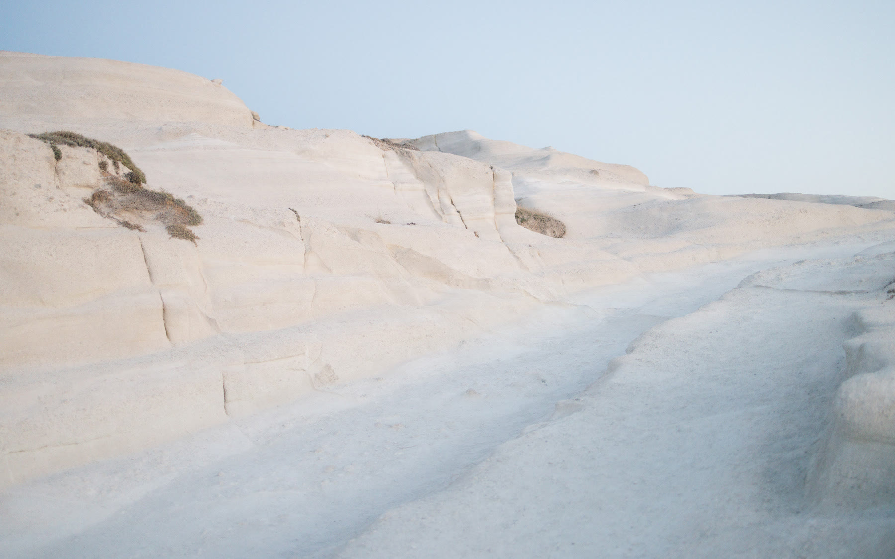 A serene coastal scene on the Greek Island of Milos features a minimalist setup with a sheet and pillow on a rocky shoreline overlooking calm waters and distant islands.